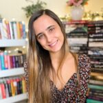 A smiling woman with long brown hair in a floral dress stands in front of a bookshelf filled with colorful books, with string lights and a vase of flowers in the background.