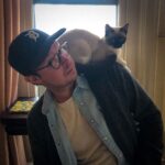 A man wearing a Detroit Tigers cap and glasses looks lovingly at a Siamese cat perched on his shoulder inside a cozy room.