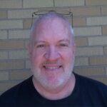 A smiling bald man with a beard, wearing glasses atop his head, posing in front of a brick wall. He is dressed in a black T-shirt.