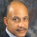 Close-up portrait of a middle-aged balding man with a mustache, wearing a dark suit and striped shirt, against a mottled brown backdrop.