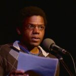 A man wearing glasses and a striped sweater over a collared shirt speaks into a microphone, holding papers in a dimly lit setting.