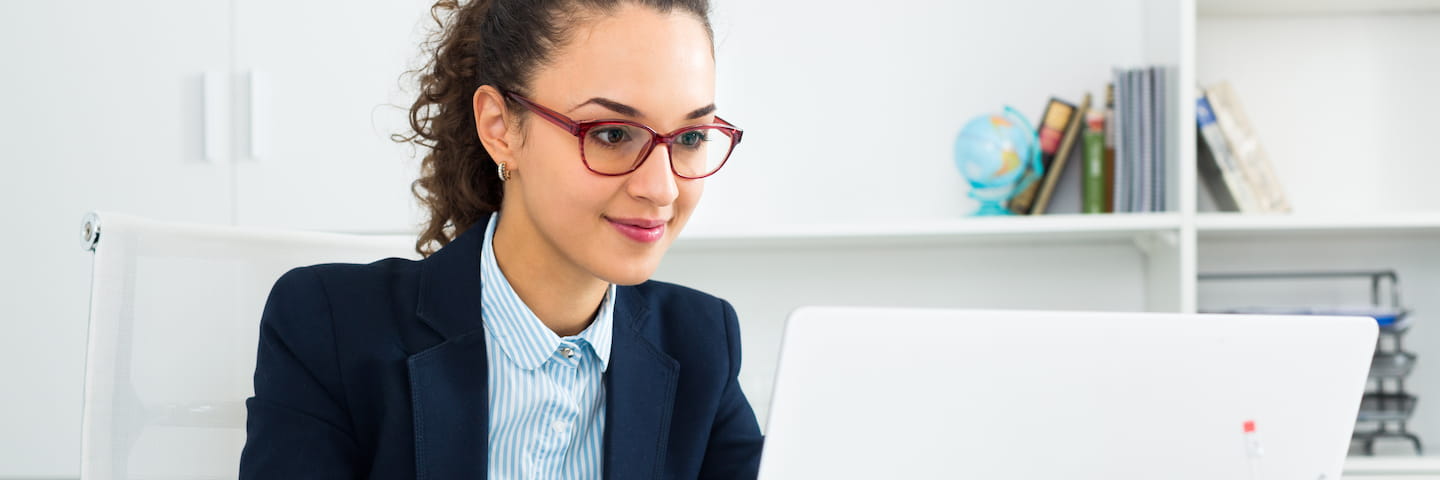 A professional woman with curly hair and glasses is working intently on a laptop in a bright office environment, pursuing her Online Business Administration degree.