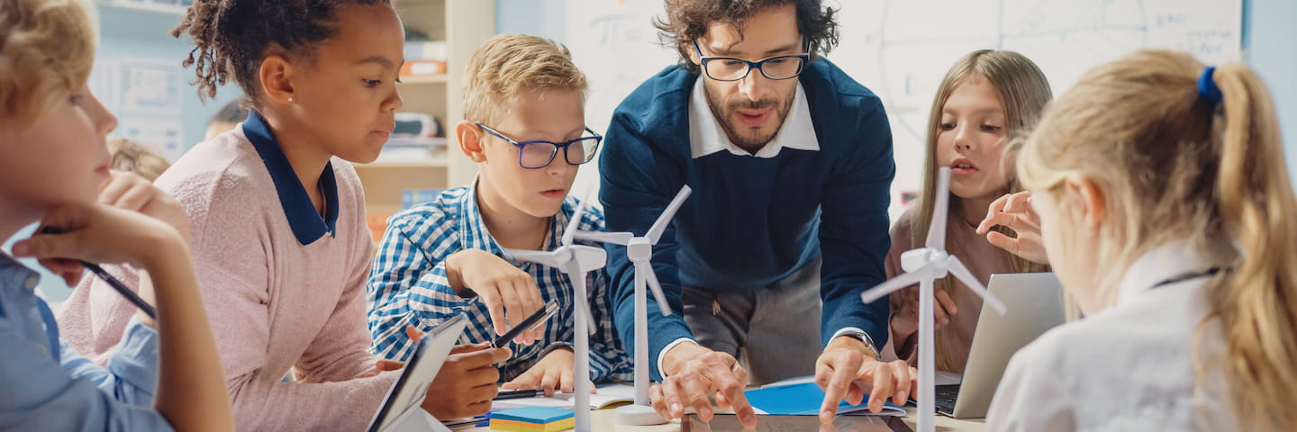 A teacher and diverse group of students engaging in an online education project with small wind turbines on a table, emphasizing teamwork and renewable energy education.