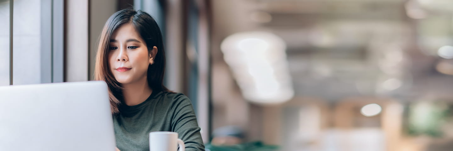 A focused woman with long black hair researches accredited online colleges on a laptop at a table near a window, with a white mug beside her, in a brightly lit, modern setting.