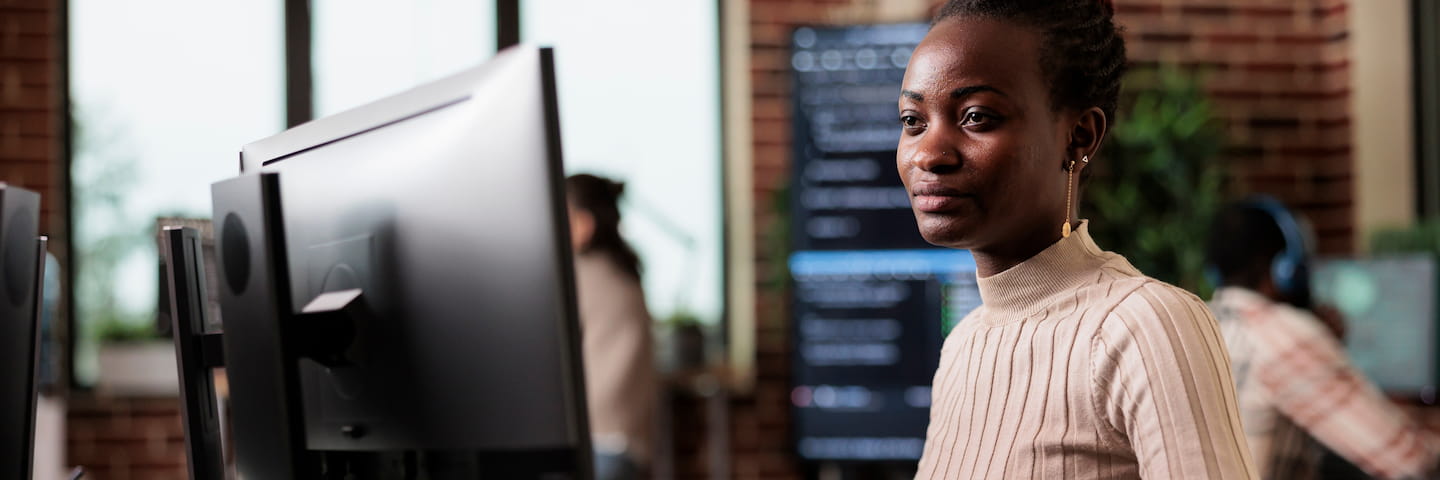 A woman in a beige top works at a dual monitor setup in a modern office, turning to look at the camera with a focused expression as she follows an online college tech guide.
