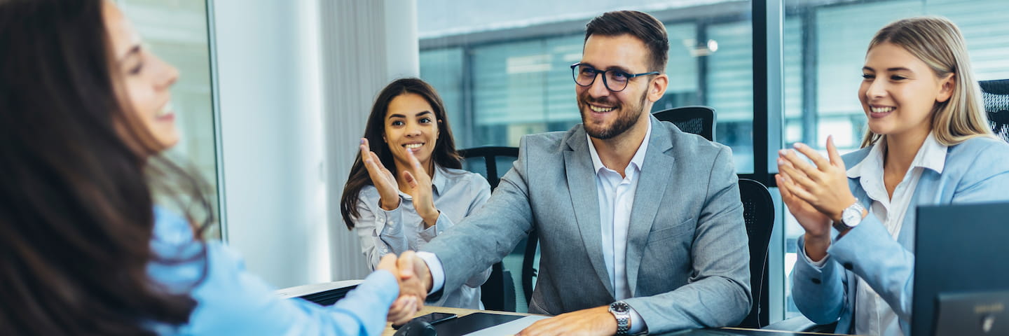 Four professionals in a modern office setting, three women and one man, smiling and clapping hands as two of them conclude a handshake after discussing the best online human resources degrees.