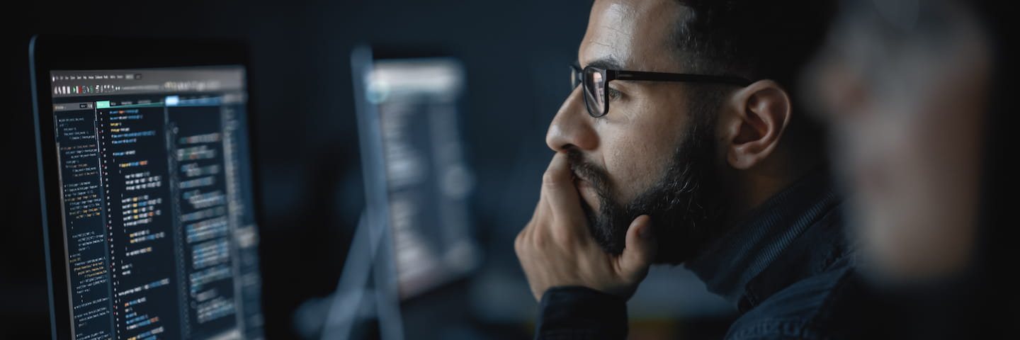 A focused man with glasses and a beard examines code on a computer screen in a dimly lit office environment, possibly enrolled in one of the top online master's programs for software engineering.