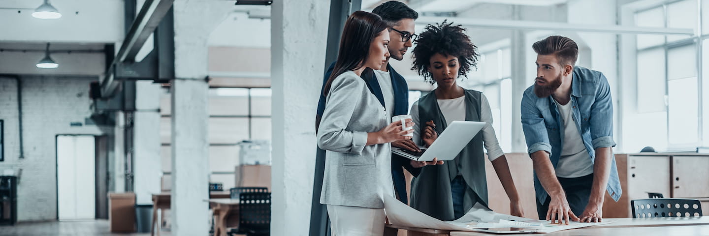 Four professionals, a diverse group of two men and two women enrolled in one of the best online MBA programs, are engaged in a discussion around a laptop in a modern office setting.