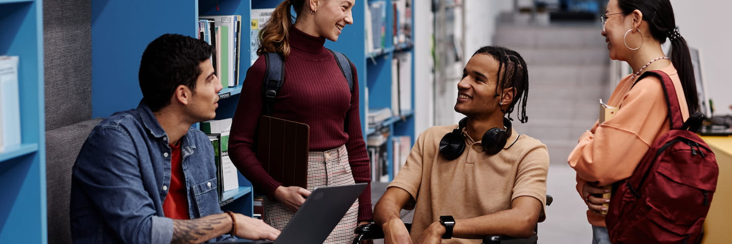 Four diverse university students, including college students with disabilities, engaging in a friendly conversation in a library aisle, with books and laptops visible.