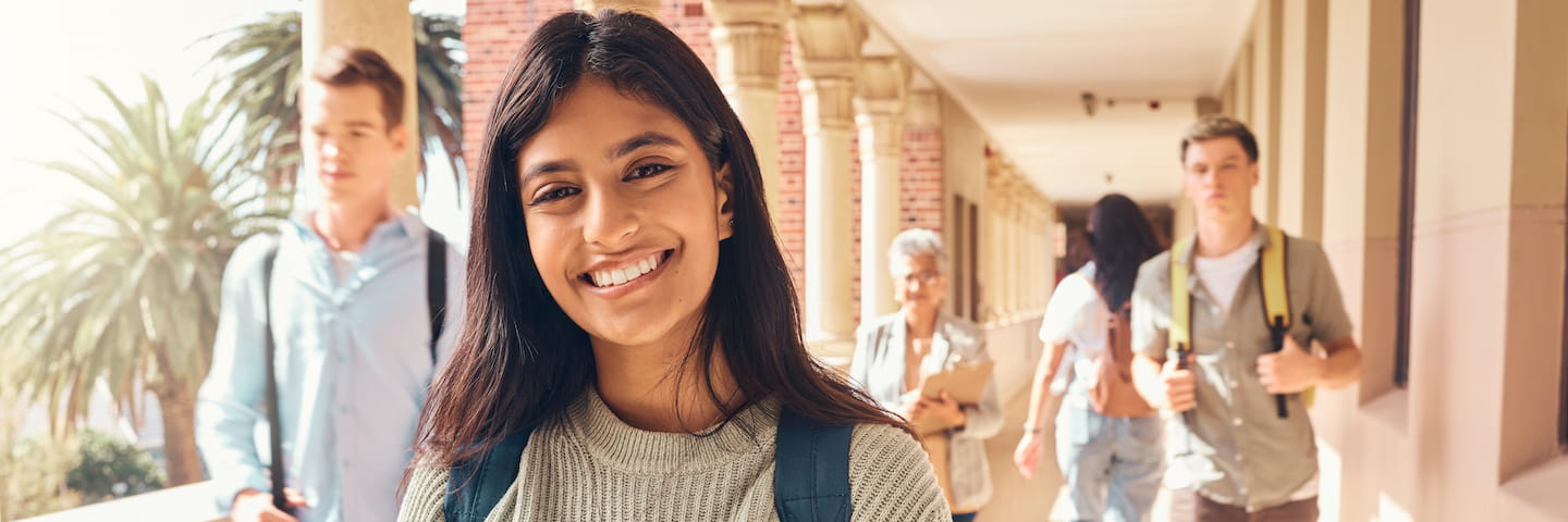 A young south Asian female college student smiling at the camera, standing in a sunlit school hallway with other students walking in the background.