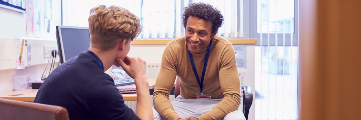 A smiling counselor with curly hair listens attentively to a young male student in an office environment, depicting a supportive session facilitated by someone with one of the best social work degrees.