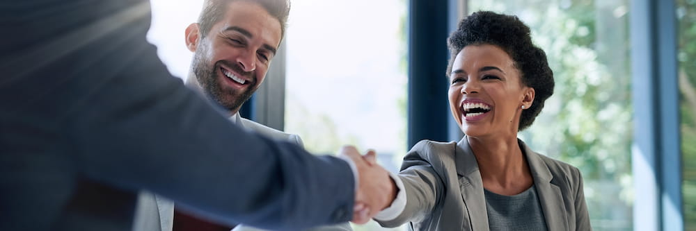 A smiling woman with curly hair shakes hands with a man in an office setting, expressing enthusiasm and congratulations in a professional environment.