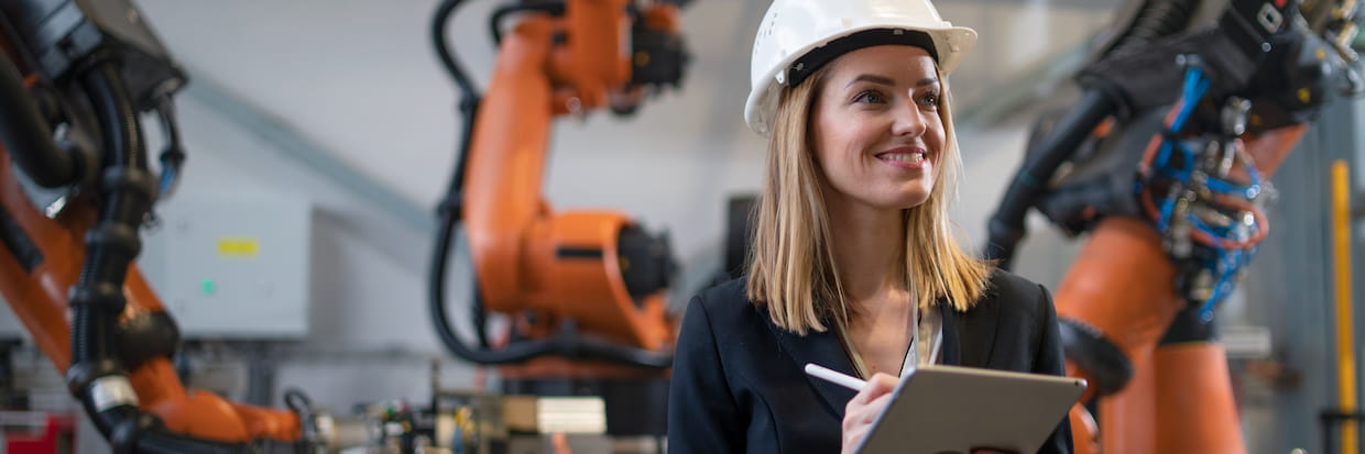 A smiling female engineer wearing a white helmet carries a tablet, standing in an industrial setting with an orange robotic arm in the background, embodying a guide to learn how to become an engineer.